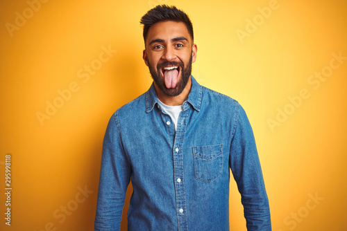 Young indian man wearing denim shirt standing over isolated yellow background sticking tongue out happy with funny expression. Emotion concept.