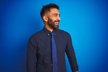 Young indian businessman wearing elegant shirt and tie standing over isolated blue background looking away to side with smile on face, natural expression. Laughing confident.