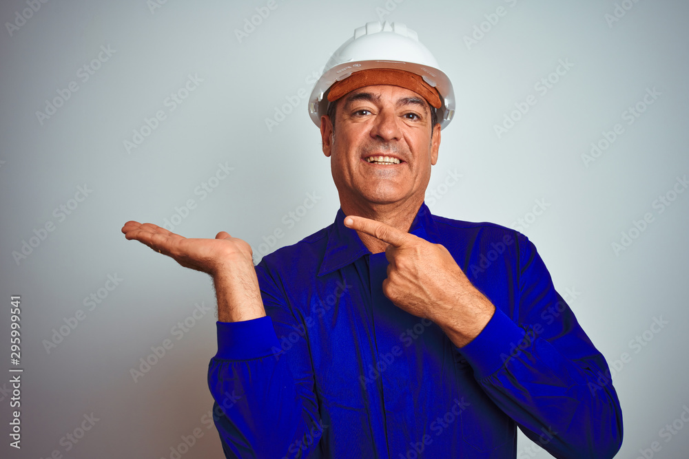 Handsome middle age worker man wearing uniform and helmet over isolated white background amazed and smiling to the camera while presenting with hand and pointing with finger.