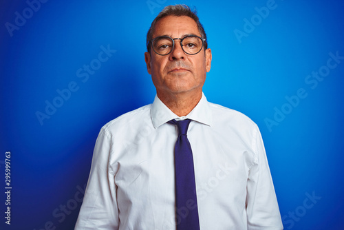 Handsome middle age businessman wearing glasses standing over isolated blue background with a confident expression on smart face thinking serious