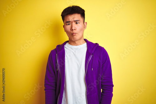 Young asian chinese man wearing purple sweatshirt standing over isolated yellow background making fish face with lips, crazy and comical gesture. Funny expression.