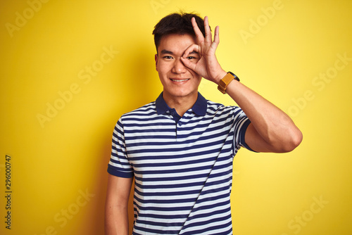 Young asian chinese man wearing striped polo standing over isolated yellow background doing ok gesture with hand smiling, eye looking through fingers with happy face.