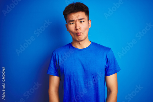 Young asian chinese man wearing t-shirt standing over isolated blue background making fish face with lips, crazy and comical gesture. Funny expression.
