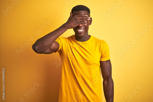 Young african american man wearing casual t-shirt standing over isolated yellow background smiling and laughing with hand on face covering eyes for surprise. Blind concept.