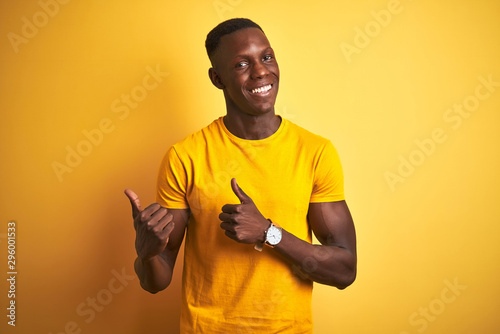 Young african american man wearing casual t-shirt standing over isolated yellow background Pointing to the back behind with hand and thumbs up, smiling confident