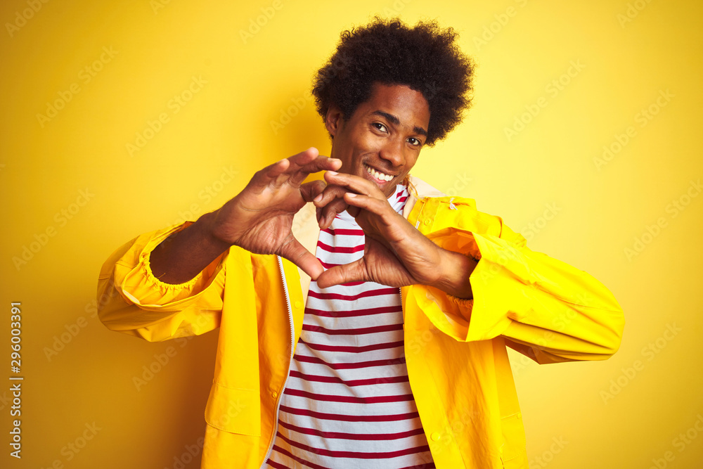 African american man with afro hair wearing rain coat standing over isolated yellow background smiling in love doing heart symbol shape with hands. Romantic concept. African american man with afro hair wearing rain coat standing over isolated yellow background smiling in love doing heart symbol shape with hands. Romantic concept.