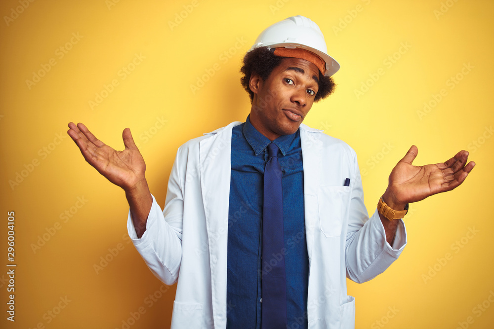 Afro american engineer man wearing white coat and helmet over isolated yellow background clueless and confused expression with arms and hands raised. Doubt concept.