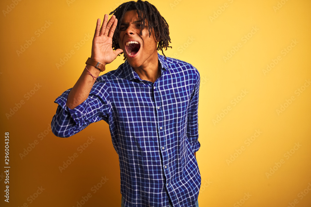 Afro man with dreadlocks wearing casual shirt standing over isolated yellow background shouting and screaming loud to side with hand on mouth. Communication concept. Afro man with dreadlocks wearing casual shirt standing over isolated yellow background shouting and screaming loud to side with hand on mouth. Communication concept.