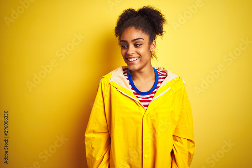 Young african american woman wearing rain coat over isolated yellow background looking away to side with smile on face, natural expression. Laughing confident.