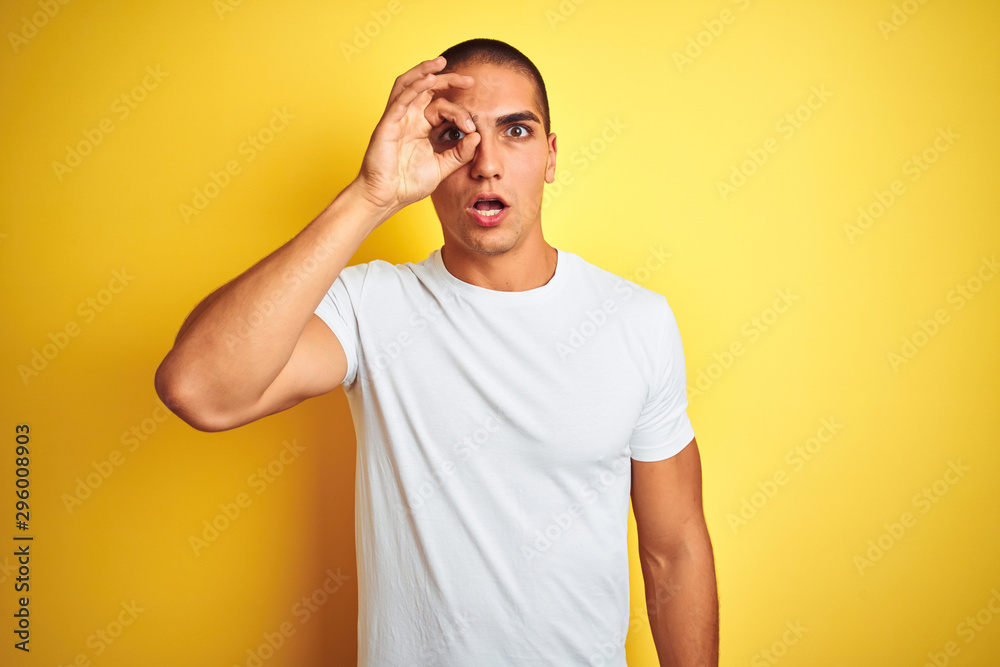 Young caucasian man wearing casual white t-shirt over yellow isolated background doing ok gesture shocked with surprised face, eye looking through fingers. Unbelieving expression. Young caucasian man wearing casual white t-shirt over yellow isolated background doing ok gesture shocked with surprised face, eye looking through fingers. Unbelieving expression.