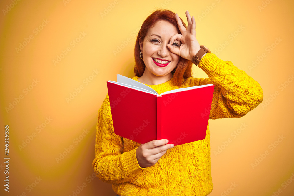 Young redhead teacher woman reading red book over yellow isolated background with happy face smiling doing ok sign with hand on eye looking through fingers Young redhead teacher woman reading red book over yellow isolated background with happy face smiling doing ok sign with hand on eye looking through fingers