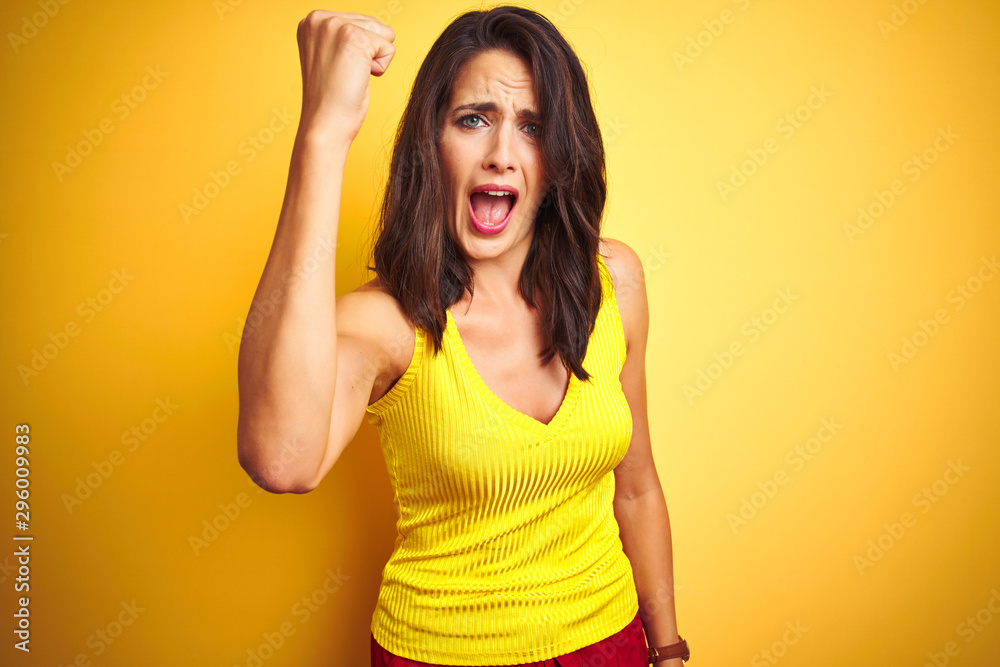 Young beautiful woman wearing t-shirt standing over yellow isolated background angry and mad raising fist frustrated and furious while shouting with anger. Rage and aggressive concept.