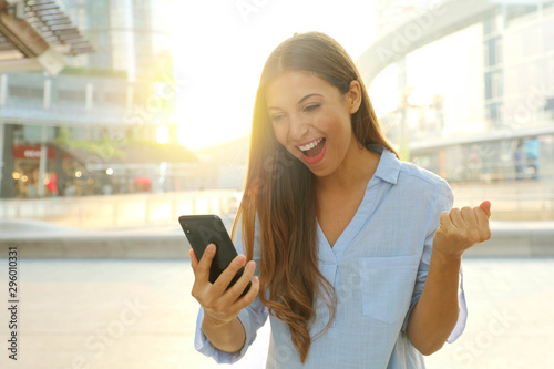 Overjoyed young woman reading message from friend on mobile expressing happiness about great news. Emotional female excited with winning discount for shopping while walking in downtown.
