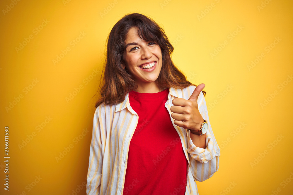 Young beautiful woman wearing red t-shirt and stripes shirt over yellow isolated background doing happy thumbs up gesture with hand. Approving expression looking at the camera showing success. Young beautiful woman wearing red t-shirt and stripes shirt over yellow isolated background doing happy thumbs up gesture with hand. Approving expression looking at the camera showing success.