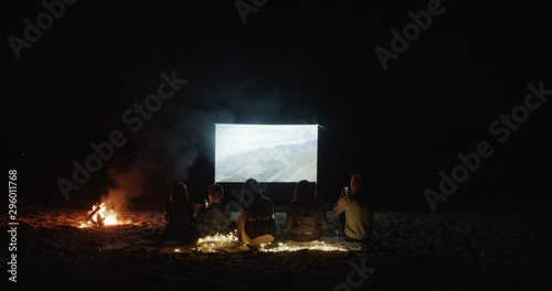 Young Friends are sitting by the fire on the beach and watching a movie with a projector by the sea. Open air cinema