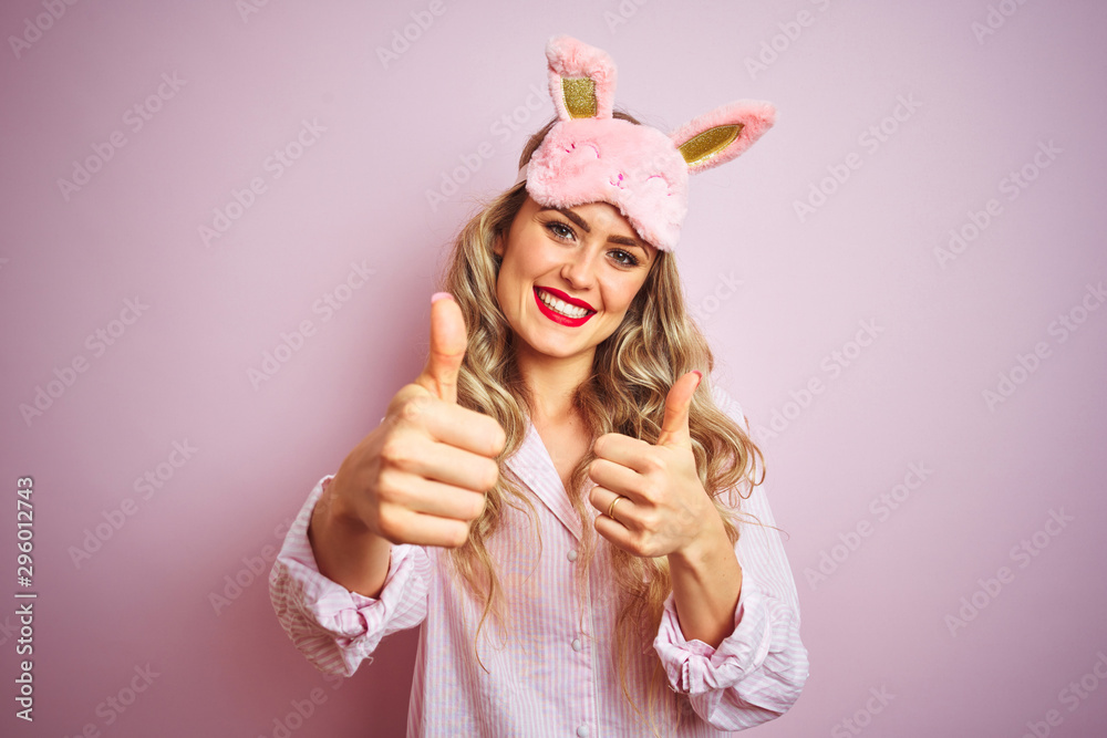 Young beautiful woman wearing pajama and sleep mask over pink isolated background approving doing positive gesture with hand, thumbs up smiling and happy for success. Winner gesture. Young beautiful woman wearing pajama and sleep mask over pink isolated background approving doing positive gesture with hand, thumbs up smiling and happy for success. Winner gesture.