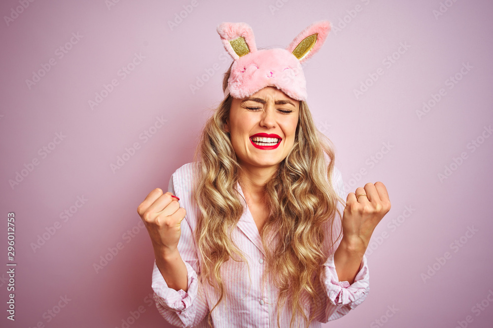Young beautiful woman wearing pajama and sleep mask over pink isolated background excited for success with arms raised and eyes closed celebrating victory smiling. Winner concept.