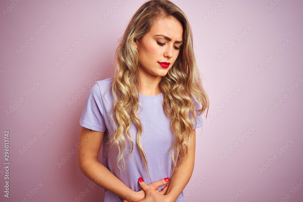 Young beautiful woman wearing purple t-shirt standing over pink isolated background with hand on stomach because nausea, painful disease feeling unwell. Ache concept. Young beautiful woman wearing purple t-shirt standing over pink isolated background with hand on stomach because nausea, painful disease feeling unwell. Ache concept.