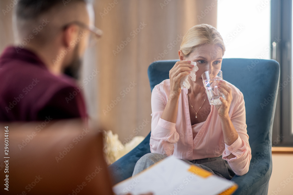 Woman drinking water and drying tears while visiting psychologist Woman drinking water and drying tears while visiting psychologist