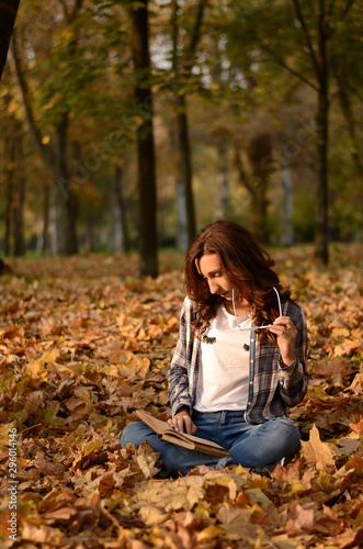 Beautiful girl with glasses sitting on the ground and reading book in park. Leisure time on warm autumn day. Autumn mood, enjoy the season