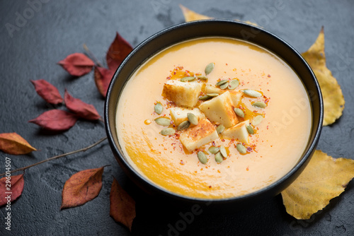 Pumpkin cream-soup with roasted cheese and pumpkin seeds, studio shot on a black stone background with autumn leaves