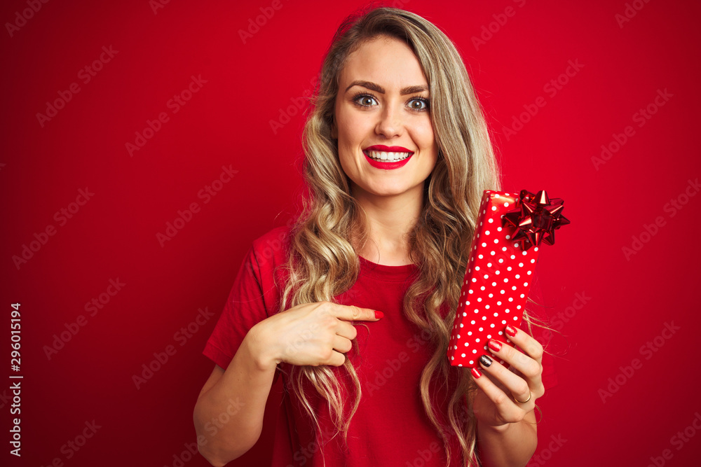 Young beautiful woman holding valentine gift over red isolated background with surprise face pointing finger to himself Young beautiful woman holding valentine gift over red isolated background with surprise face pointing finger to himself