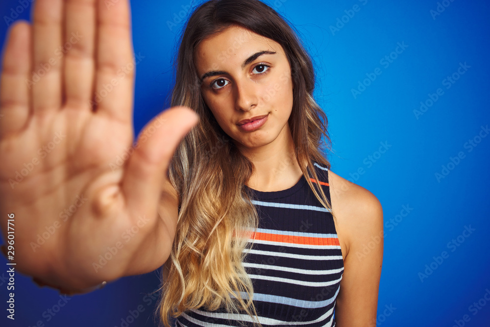 Young beautiful woman wearing stripes t-shirt over blue isolated background with open hand doing stop sign with serious and confident expression, defense gesture