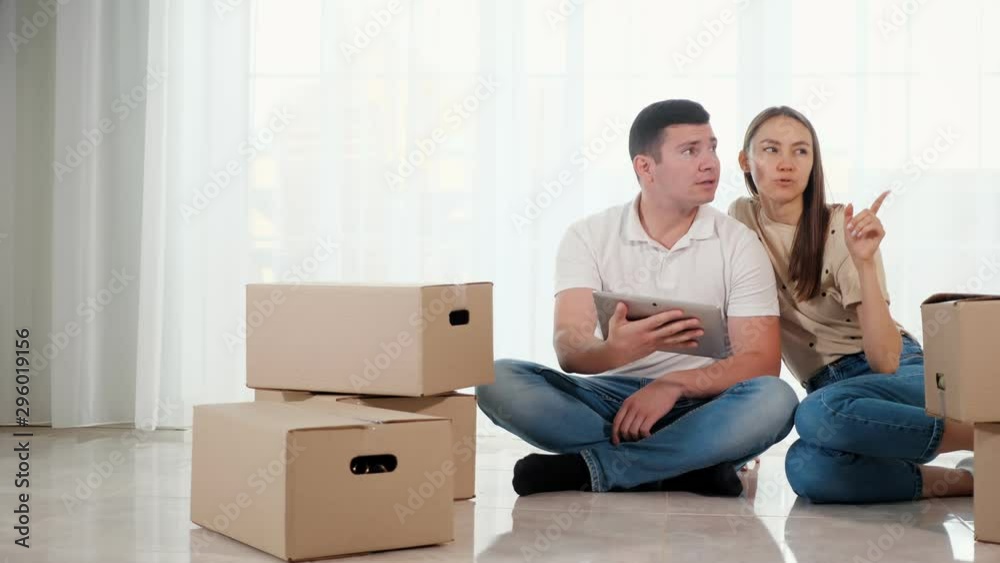 couple wife husband plans furniture location in new house sitting on floor among big cardboard boxes and uses tablet