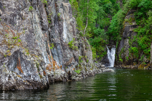 A picturesque natural place with a waterfall flowing from the Altai mountains and flowing into the Teletskoye Lake around rocky cliffs with green trees and grass. Travel and tourism in nature.