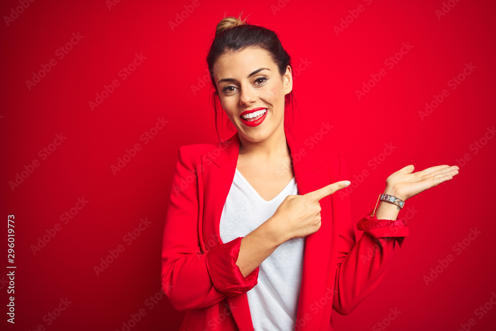 Young beautiful business woman standing over red isolated background amazed and smiling to the camera while presenting with hand and pointing with finger.