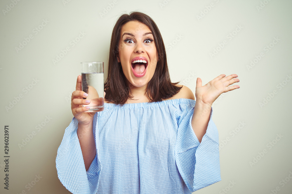 Young beautiful woman drinking a glass of fresh water over isolated background very happy and excited, winner expression celebrating victory screaming with big smile and raised hands Young beautiful woman drinking a glass of fresh water over isolated background very happy and excited, winner expression celebrating victory screaming with big smile and raised hands