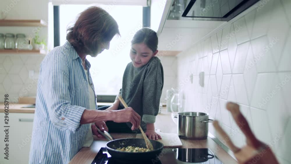A small girl with senior grandmother indoors in kitchen cooking. A small girl with senior grandmother indoors in kitchen cooking.