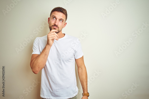 Young handsome man wearing casual white t-shirt over isolated background with hand on chin thinking about question, pensive expression. Smiling with thoughtful face. Doubt concept.