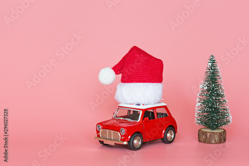 Red toy car with a santa hat, next christmas tree on pink paper background. Winter delivery, xmas, happy new year 2020 celebration concept. greeting card, mockup, copy space, place for text, template