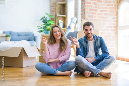 Young beautiful couple moving to a new house sitting on the floor with a big smile on face, pointing with hand and finger to the side looking at the camera. © Krakenimages.com