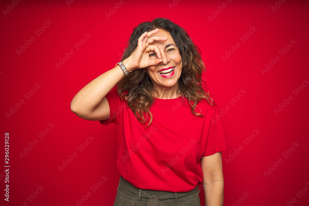 Middle age senior woman with curly hair over red isolated background doing ok gesture with hand smiling, eye looking through fingers with happy face.