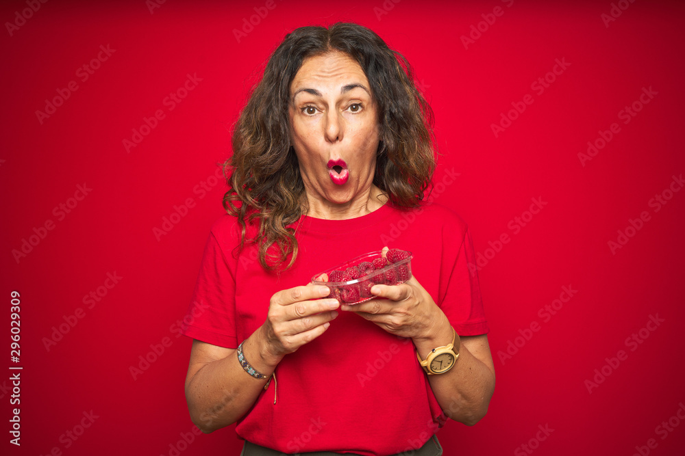Middle age senior woman eating raspberries over red isolated background scared in shock with a surprise face, afraid and excited with fear expression Middle age senior woman eating raspberries over red isolated background scared in shock with a surprise face, afraid and excited with fear expression