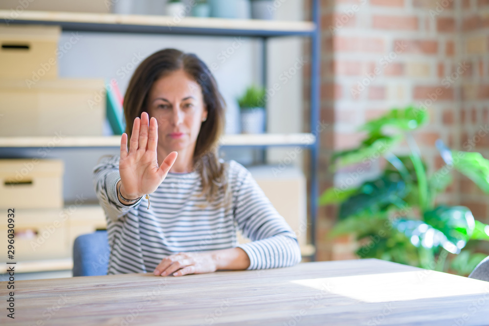 Middle age senior woman sitting at the table at home doing stop sing with palm of the hand. Warning expression with negative and serious gesture on the face. Middle age senior woman sitting at the table at home doing stop sing with palm of the hand. Warning expression with negative and serious gesture on the face.
