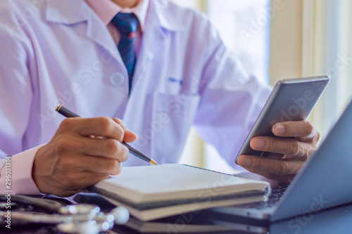 Male doctor in white lab coat,hand using mobile phone,work on laptop computer and writing note on paperwork with medical stethoscope on desk.Digital health,online medical learning,telemedicine concept