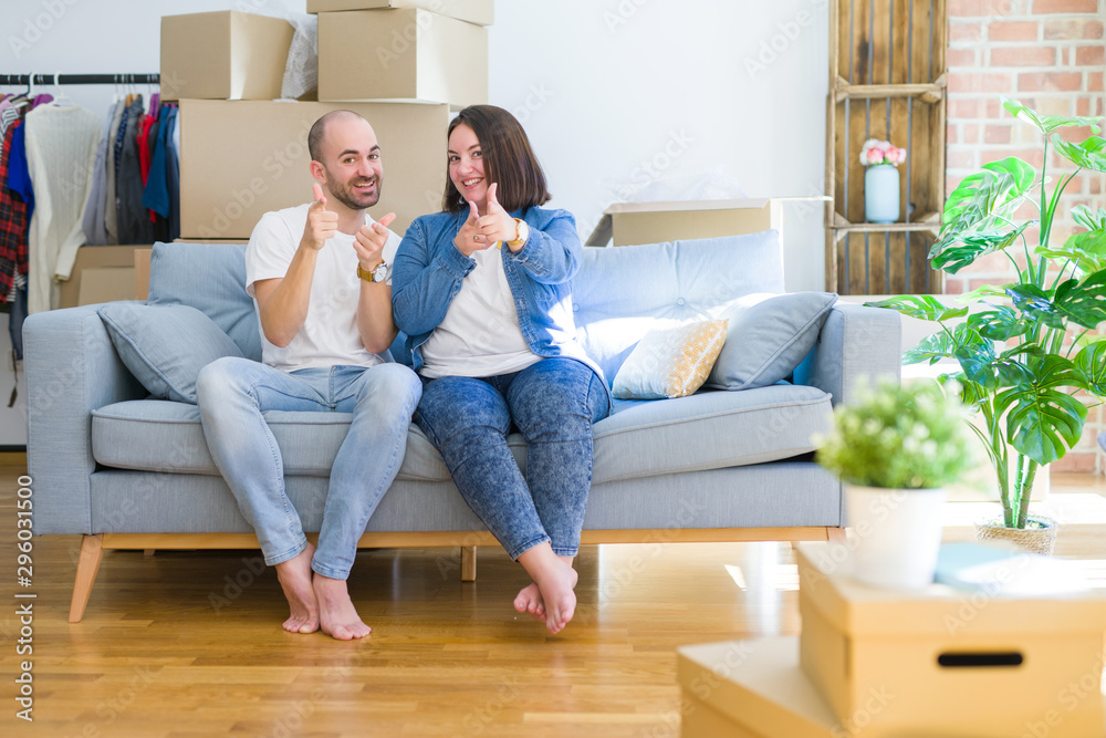 Young couple sitting on the sofa arround cardboard boxes moving to a new house pointing fingers to camera with happy and funny face. Good energy and vibes. Young couple sitting on the sofa arround cardboard boxes moving to a new house pointing fingers to camera with happy and funny face. Good energy and vibes.