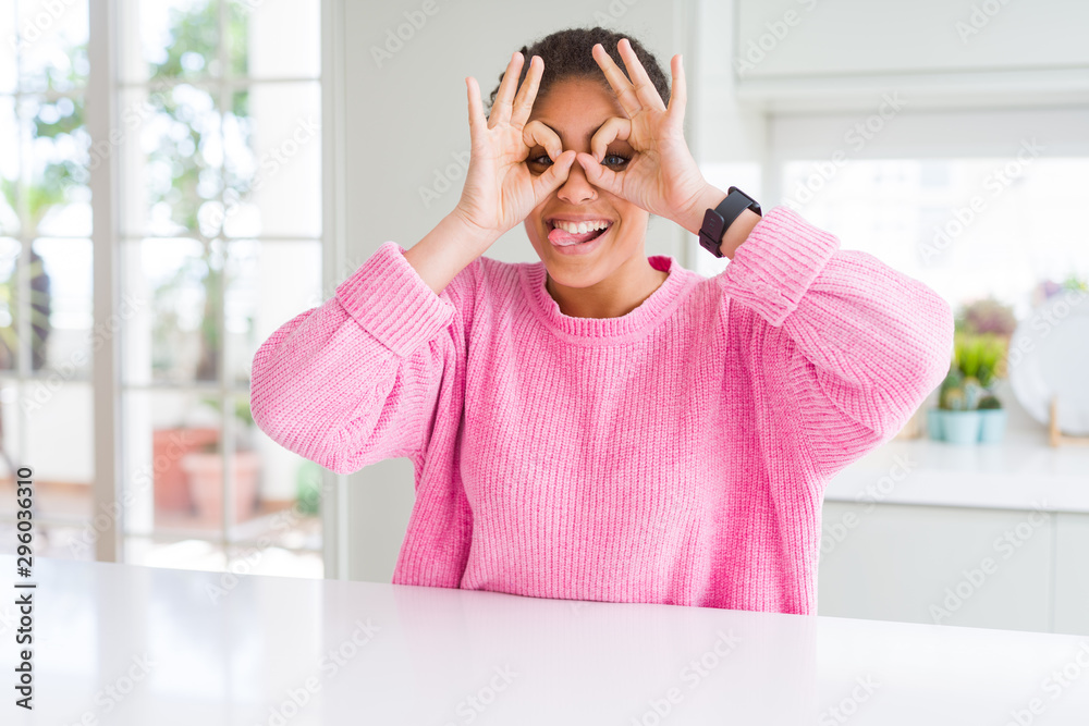 Beautiful african american woman with afro hair wearing casual pink sweater doing ok gesture like binoculars sticking tongue out, eyes looking through fingers. Crazy expression. Beautiful african american woman with afro hair wearing casual pink sweater doing ok gesture like binoculars sticking tongue out, eyes looking through fingers. Crazy expression.