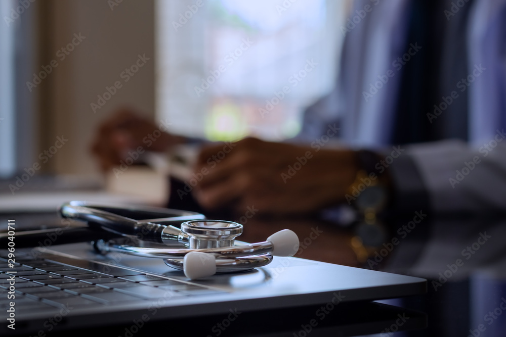Closeup image of medical stethoscope  on modern computer notebook with male doctor reading textbook blurred background. Online medical, medic tech concept. 