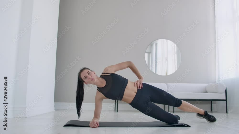 The brunette in the apartment does an exercise lateral plank standing on her knee and raising her leg up. Fitness marathon