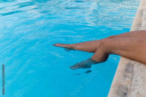 Beautiful woman legs in swimming pool. Tanned feet with pedicure in the pool water. Close-up portrait of tanned female slender legs in a warm blue pool on vacation