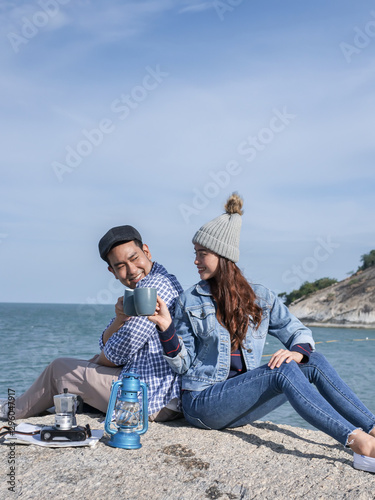 Happy Asian couple holding coffee cups on mountain peak together.