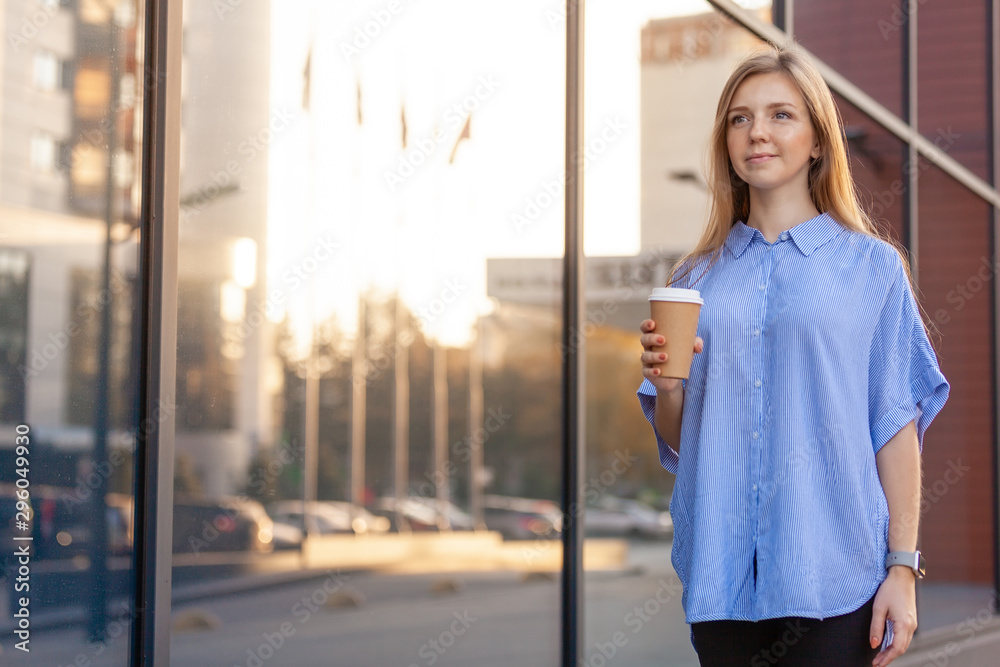 Happy cute young woman with long hair holding a coffee to go in disposable paper cup, walking along the windows of office building.