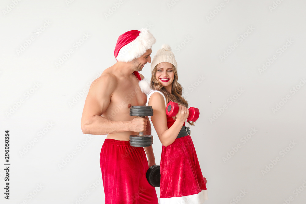 Obraz premium Young couple dressed as Santa Claus with dumbbells on light background