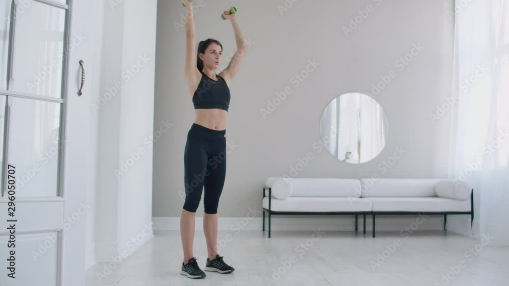 Home exercises for a beautiful body, a young brunette woman lifts dumbbells in her arms up in slow motion Home exercises for a beautiful body, a young brunette woman lifts dumbbells in her arms up in slow motion
