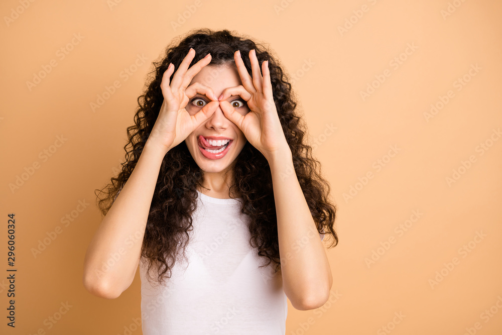 Fototapeta premium Closeup photo of funny lady playful mood holding round fingers in specs shape near eyes sticking tongue wear white casual outfit isolated beige pastel color background