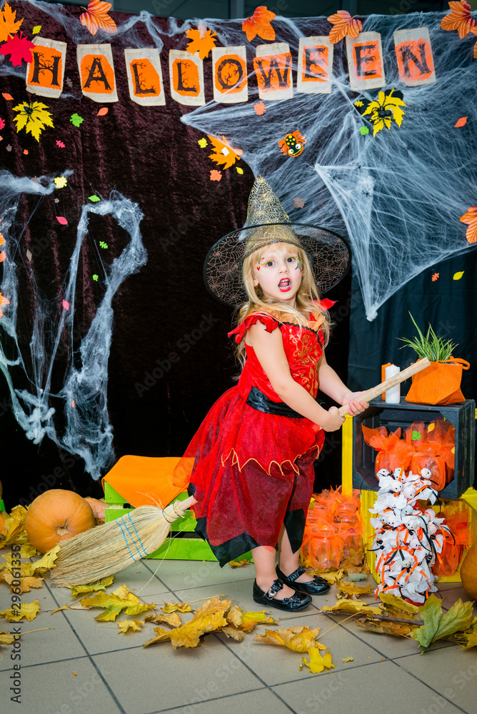 Fototapeta premium A child, a little girl in the shape of a witch on a broomstick, poses against the backdrop of scenery of cobwebs, pumpkins and autumn leaves on a Halloween holiday.